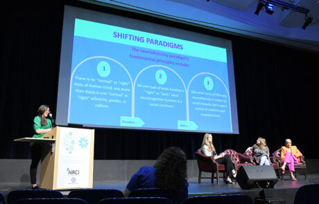 woman with glasses and green outfit speaking on stage behind a podium with a slide titled shifting paradigms on screen
