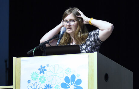 with yellow wristwatch and glasses speaking on stage behind a podium
