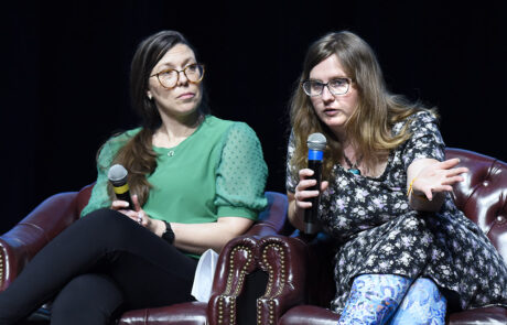 two women in dark red chairs speaking while holding microphones